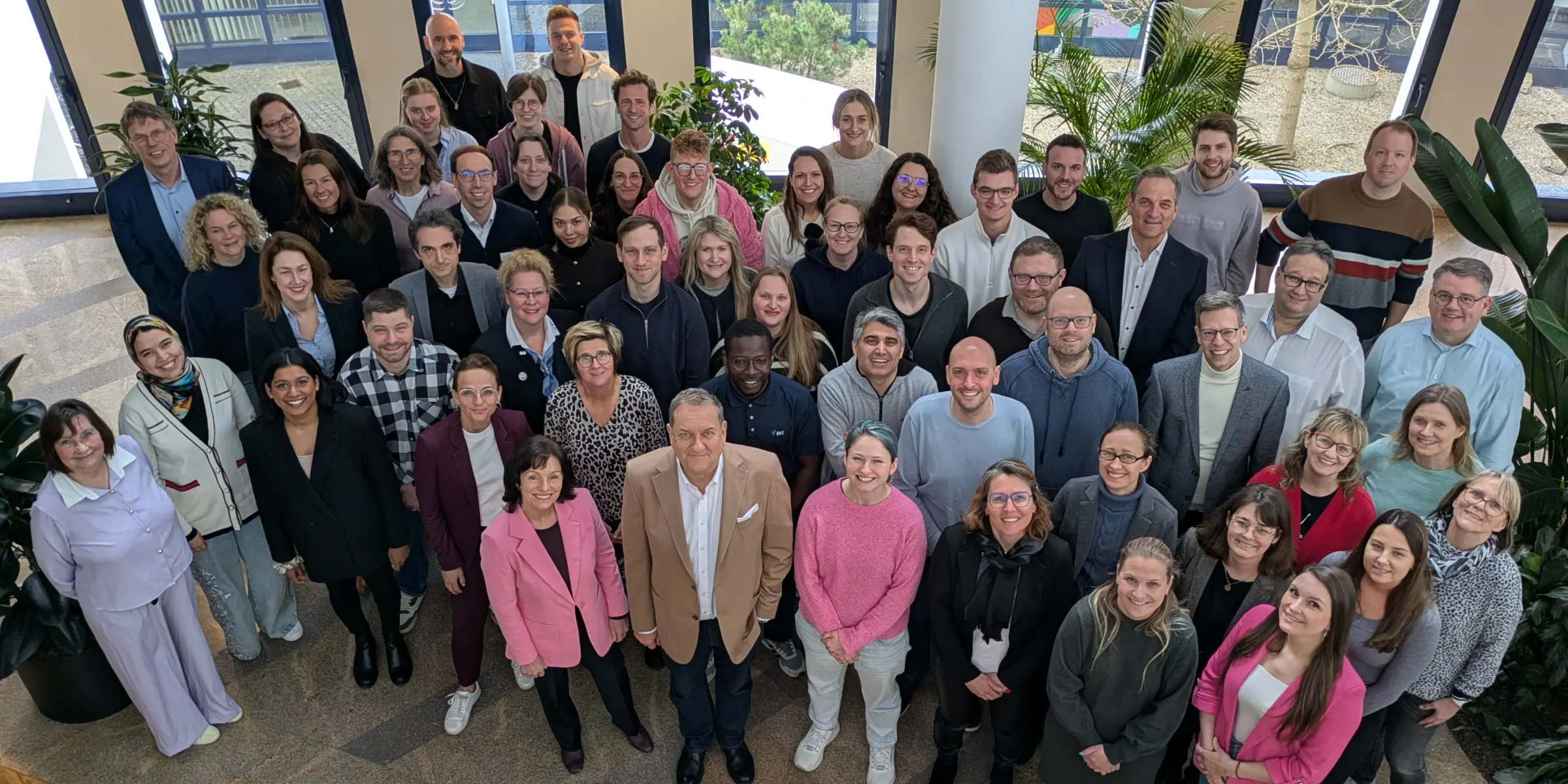 Eine Gruppe von 57 lächelnden Menschen stehen hier stellvertretend für das gesamte BVS-Team. Die Personen sind unterschiedlich gekleidet: von pink bis schwarz und von leger bis repräsentativ. Sie stehen in einer hellen Aula mit Pflanzen.