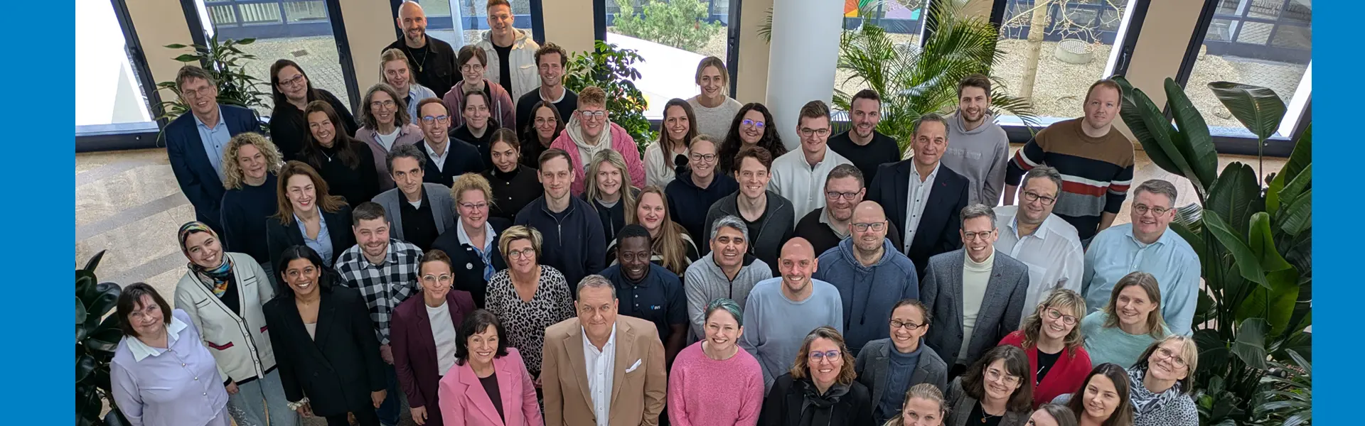 Eine Gruppe von 57 lächelnden Menschen stehen hier stellvertretend für das gesamte BVS-Team. Die Personen sind unterschiedlich gekleidet: von pink bis schwarz und von leger bis repräsentativ. Sie stehen in einer hellen Aula mit Pflanzen.